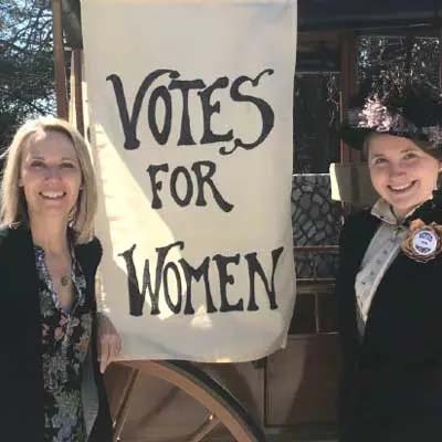 a professor and a student in suffrage era costuming pose with a banner that reads 'votes for women'