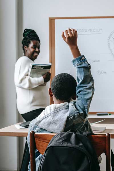 a teacher smiling at a young student raising his hand in a classroom